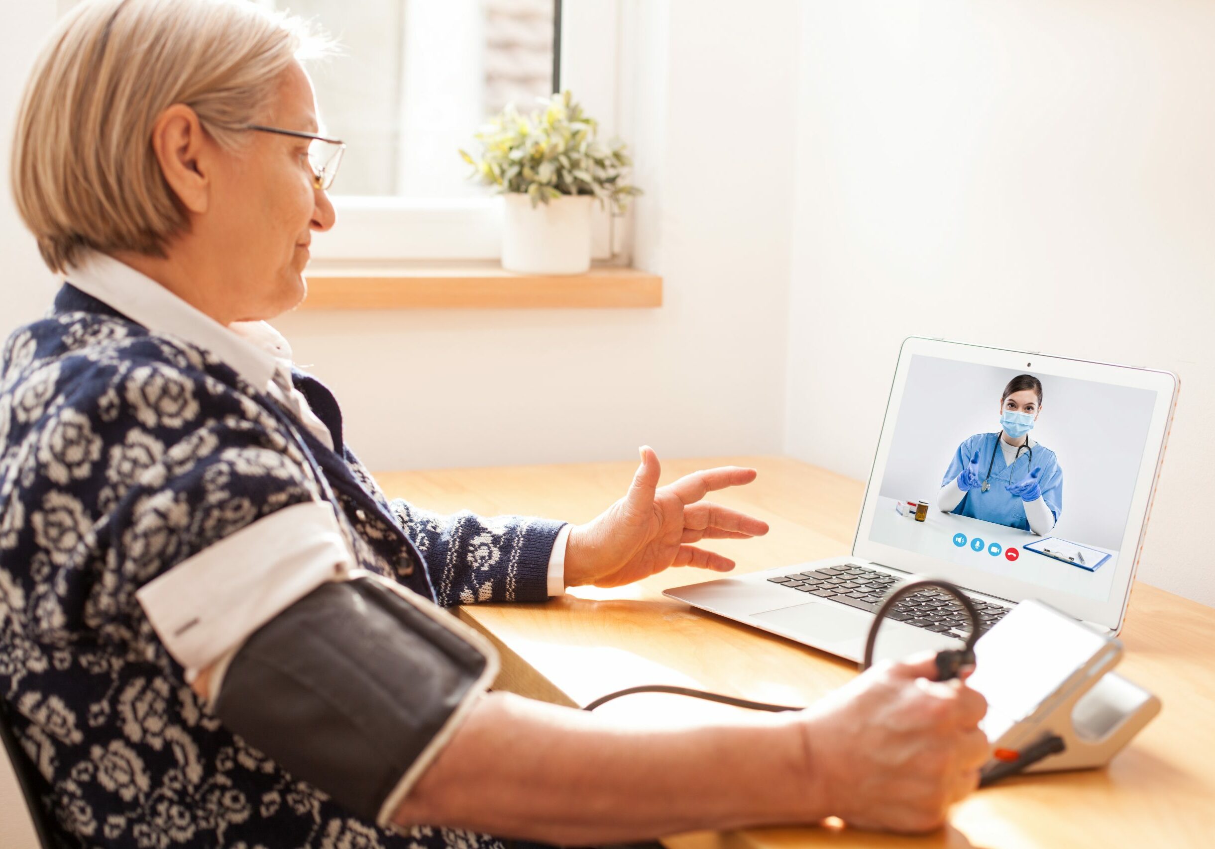 Elderly senior retired woman using sphygmomanometer blood pressure monitor to measure heart rate pulse,talking to female e-doctor via online video call help line,self-monitoring remote telemedicine